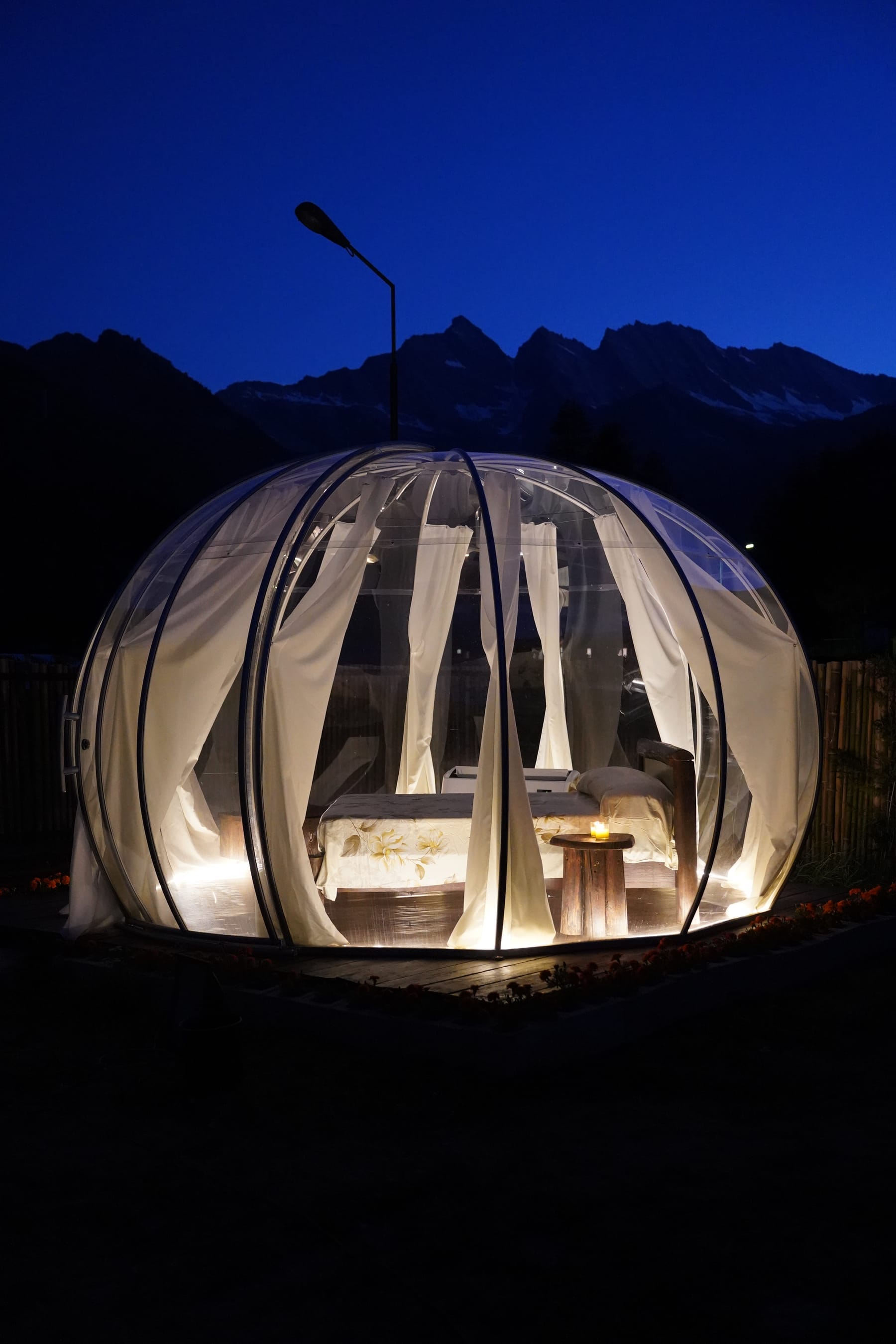 Cupola glamping trasparente illuminata di notte davanti alle Alpi